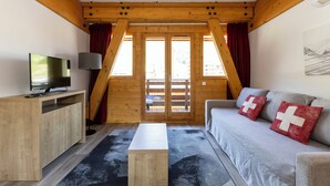 Ein Zimmer mit Holzboden, einem Fernseher auf einem Holztischchen, einer Couch mit roten Kissen und Blick durch eine Tür.