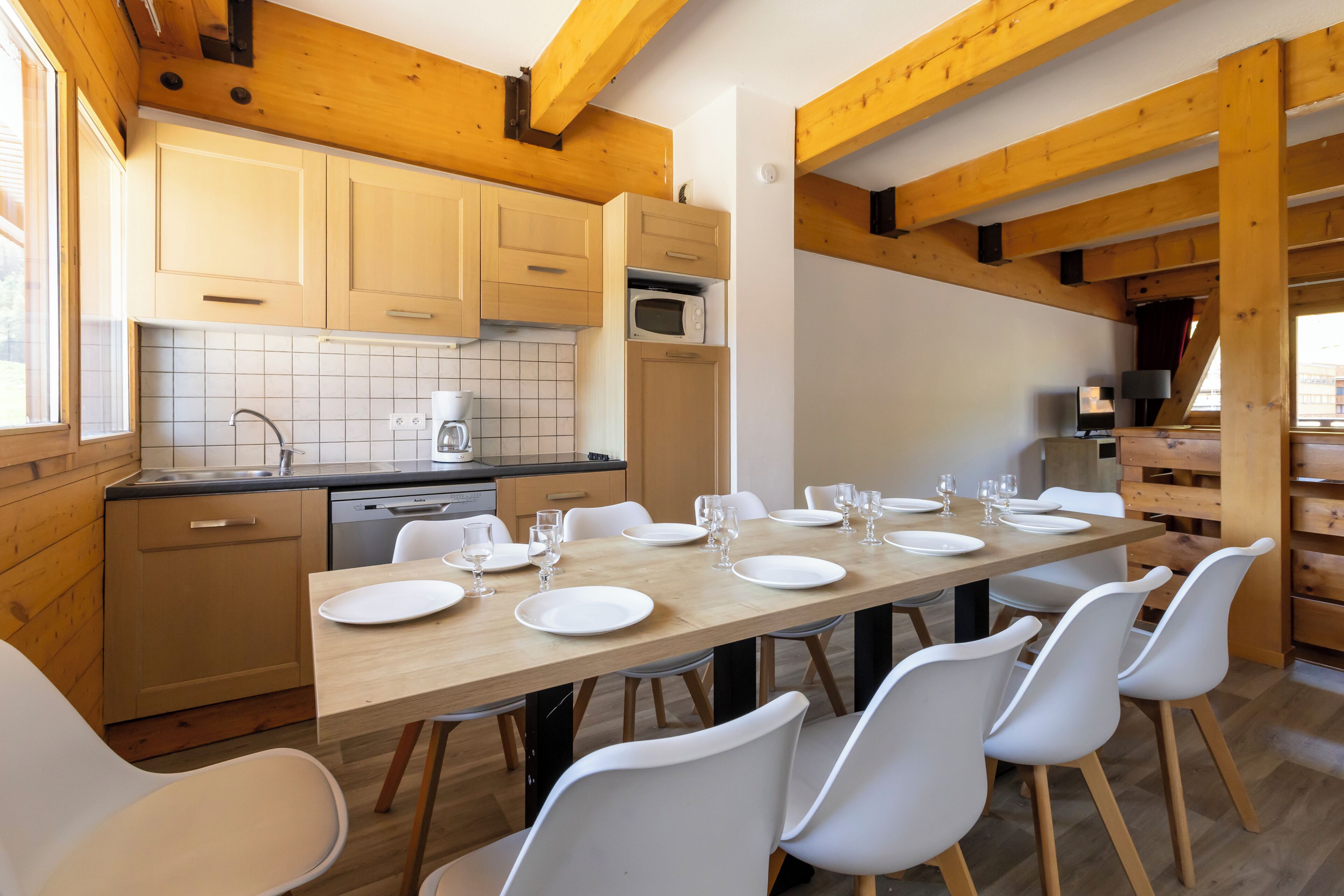 Une cuisine moderne avec des poutres en bois, une table à manger dressée pour six personnes et des chaises blanches.
