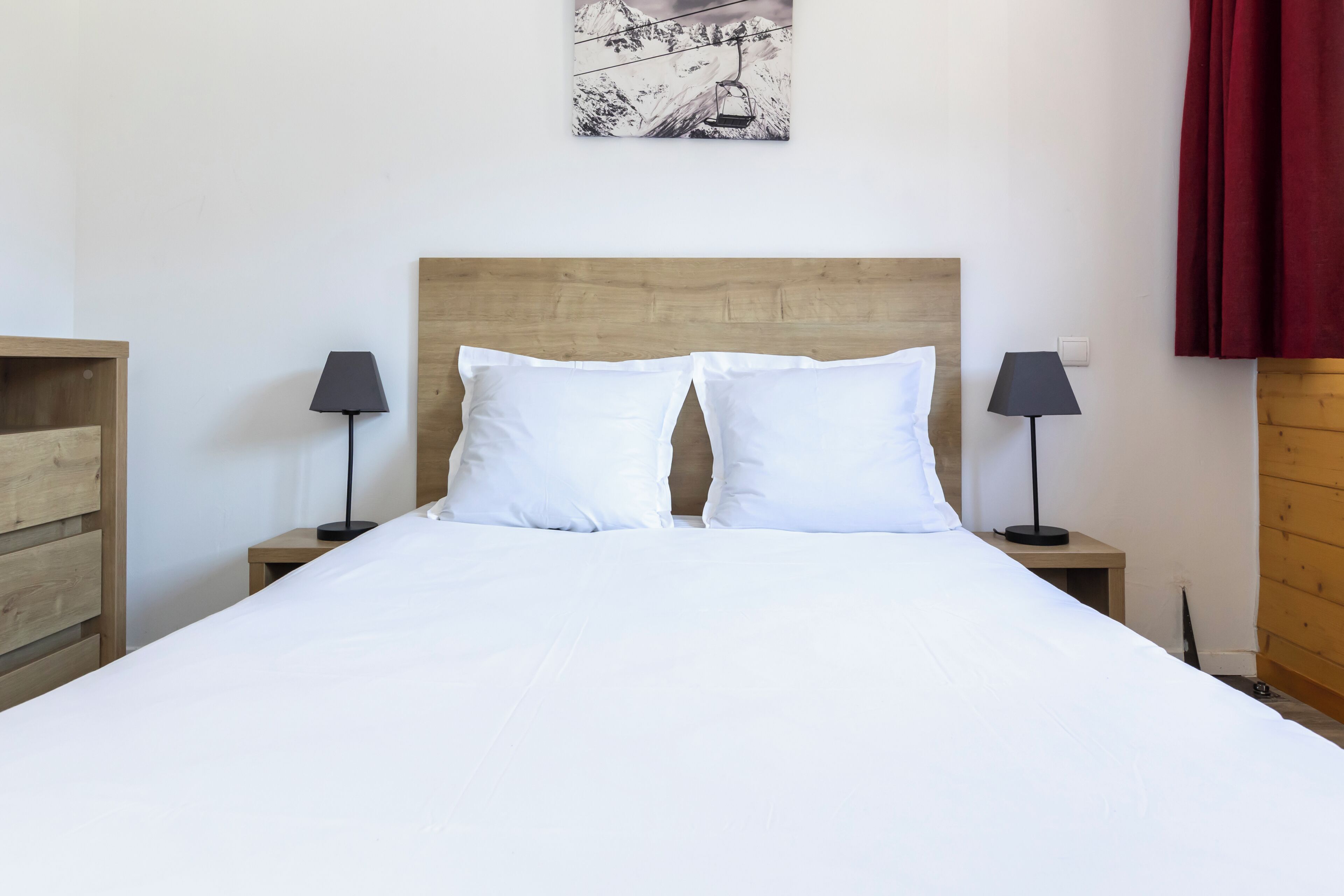 A neatly made bed with white linens, two bedside tables with lamps, a wooden headboard, and a wall-mounted picture.