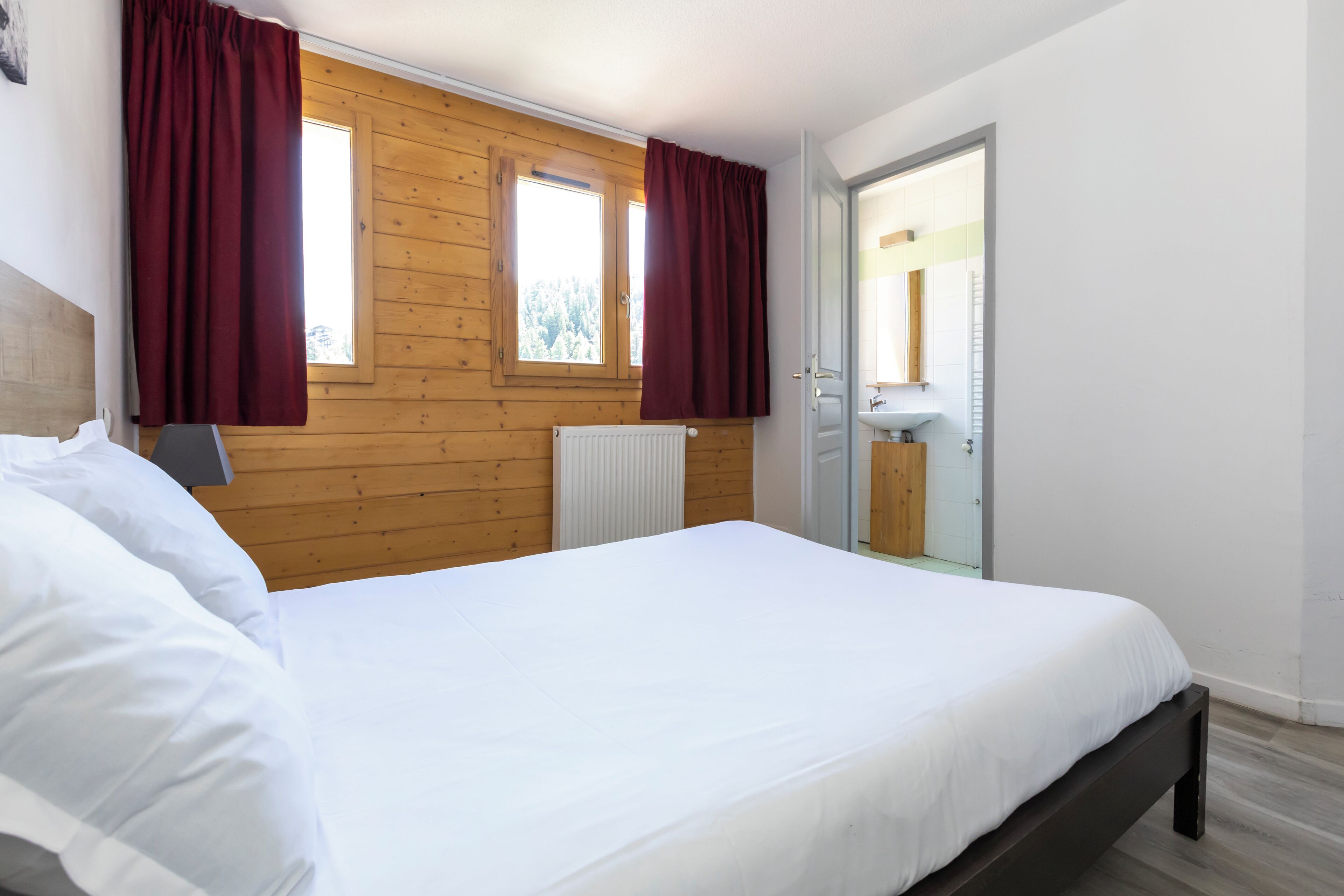 A bedroom with a wooden paneled wall, a bed with white bedding, a radiator, and a window with curtains.