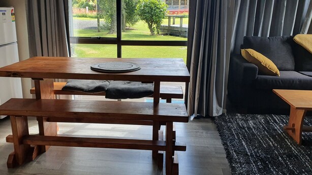 Une table en bois avec un set de table et une serviette pliée, un banc, un canapé avec des coussins et une vue sur un jardin par une fenêtre ornée de rideaux.