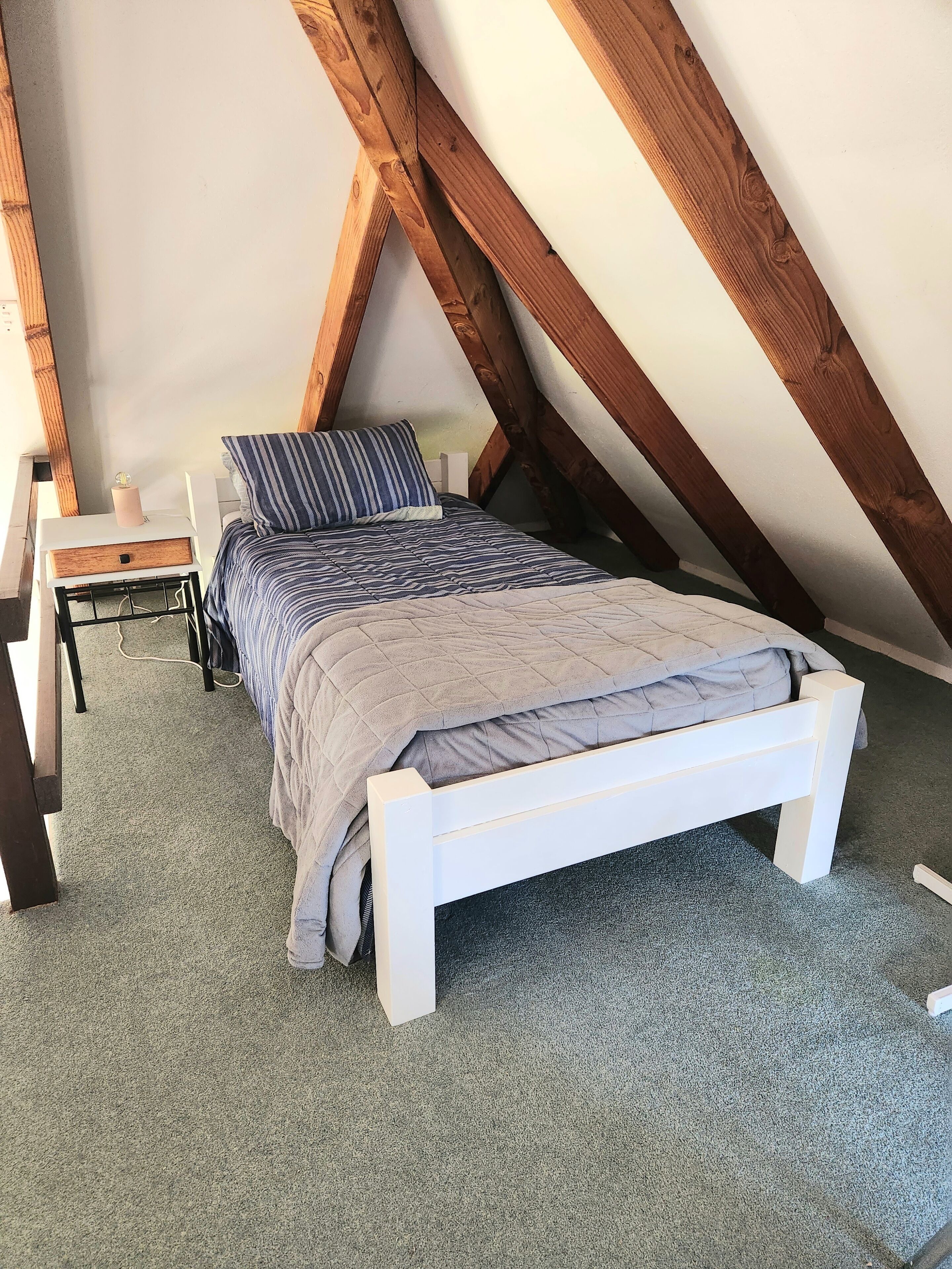 Un dormitorio con una cama blanca, una mesita de noche y una silla. La habitación tiene un techo inclinado con vigas de madera.