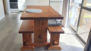 Une table de salle à manger en bois, accompagnée d’un banc, d’un set de table en fibres tressées et offrant une vue sur une terrasse à travers une porte vitrée.