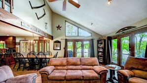 A spacious living room with a large brown leather sofa, dining area with wooden chairs, a chandelier, and large windows offering a view of greenery.