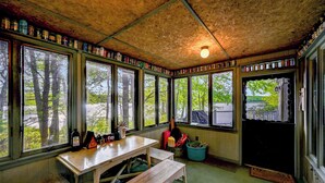 A rustic cabin with large windows, a wooden table, and shelves with jars.