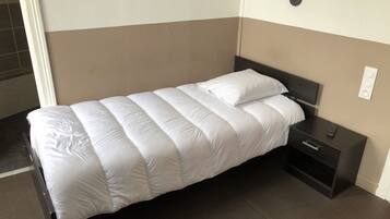 A single bed with a white duvet and pillows, a dark headboard, a bedside table with a remote control, and a wall-mounted light fixture.