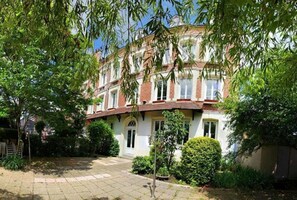 A traditional two-story building with a brick facade, a tiled courtyard, and mature trees. - Hôtel de France (Lillebonne)