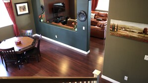 A dining area with a wooden table and chairs, a television mounted on a curved wall, a brown leather sofa, and a painting on the wall. - Best Kept Secret! 4 King Beds ·Hot Tub · 9ft Pool Table · 5 Min to South Point (Las Vegas)
