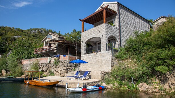 A stone house with a balcony, a dock with boats, and a small waterfall.