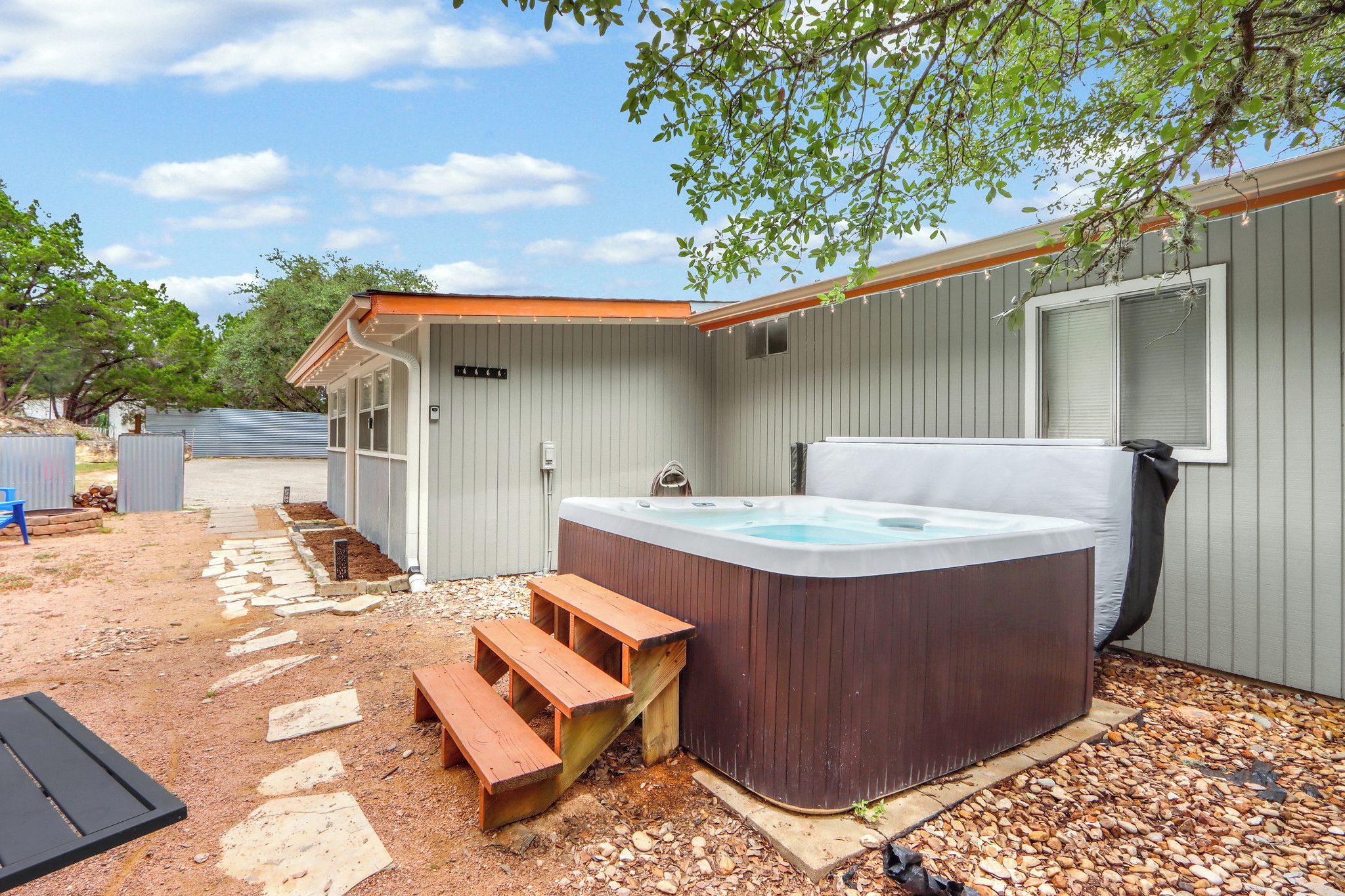 A hot tub with wooden steps, a covered patio, and a metal fence in a backyard setting.