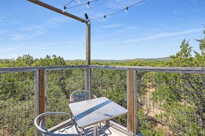 A balcony with a metal table and chair, overlooking a fenced garden with trees and a clear sky. - Come On Inn | Close to Downtown Wimberley! (Wimberley)