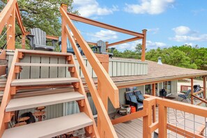 Una terraza de madera con una escalera que lleva a una zona cubierta, con sillas Adirondack y una parrilla.
