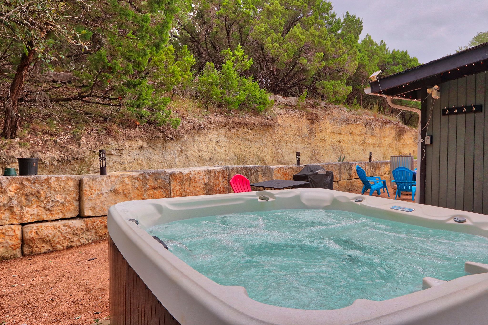 A hot tub with a stone wall backdrop, outdoor seating, and trees.