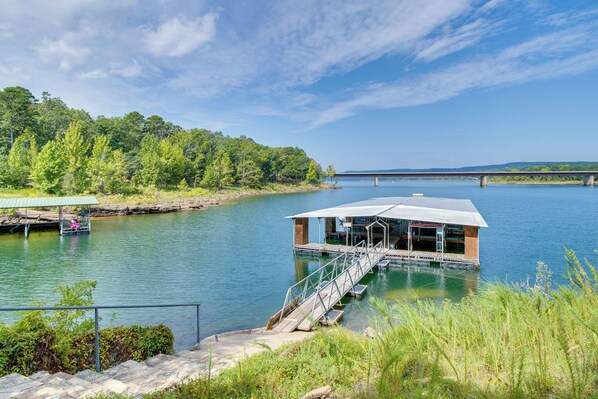 Une cabane au bord de la rivière, dotée d’un quai, entourée de verdure et avec un pont en arrière-plan.
