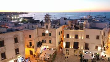 Una piazza storica con una torre dell'orologio imponente, circondata da edifici con balconi e aree con tavoli all'aperto.
