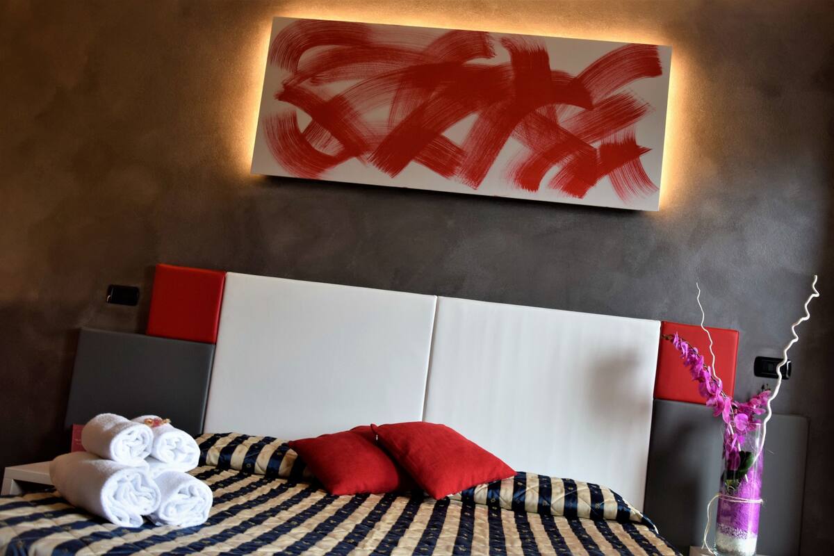 Ein Hotelzimmer mit einem großen Bett, zwei roten Kissen, einer gestreiften Bettdecke, einem an der Wand hängenden abstrakten Gemälde, einer Vase mit Blumen und einem Nachttisch mit gerollten Handtüchern.