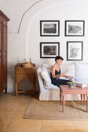 Ein Zimmer mit einem weißen Sofa, einem Holztisch und einem Korbtisch mit Küchenutensilien. An der Wand befinden sich gerahmte Bilder.