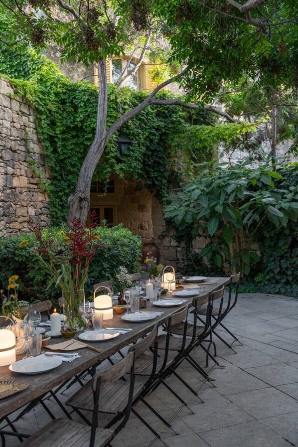 An outdoor dining area with a long wooden table set for a meal, surrounded by lush greenery and a stone wall. - Luxurious Grand 18th C. Palace with Gardens & Pool (Ghaxaq)