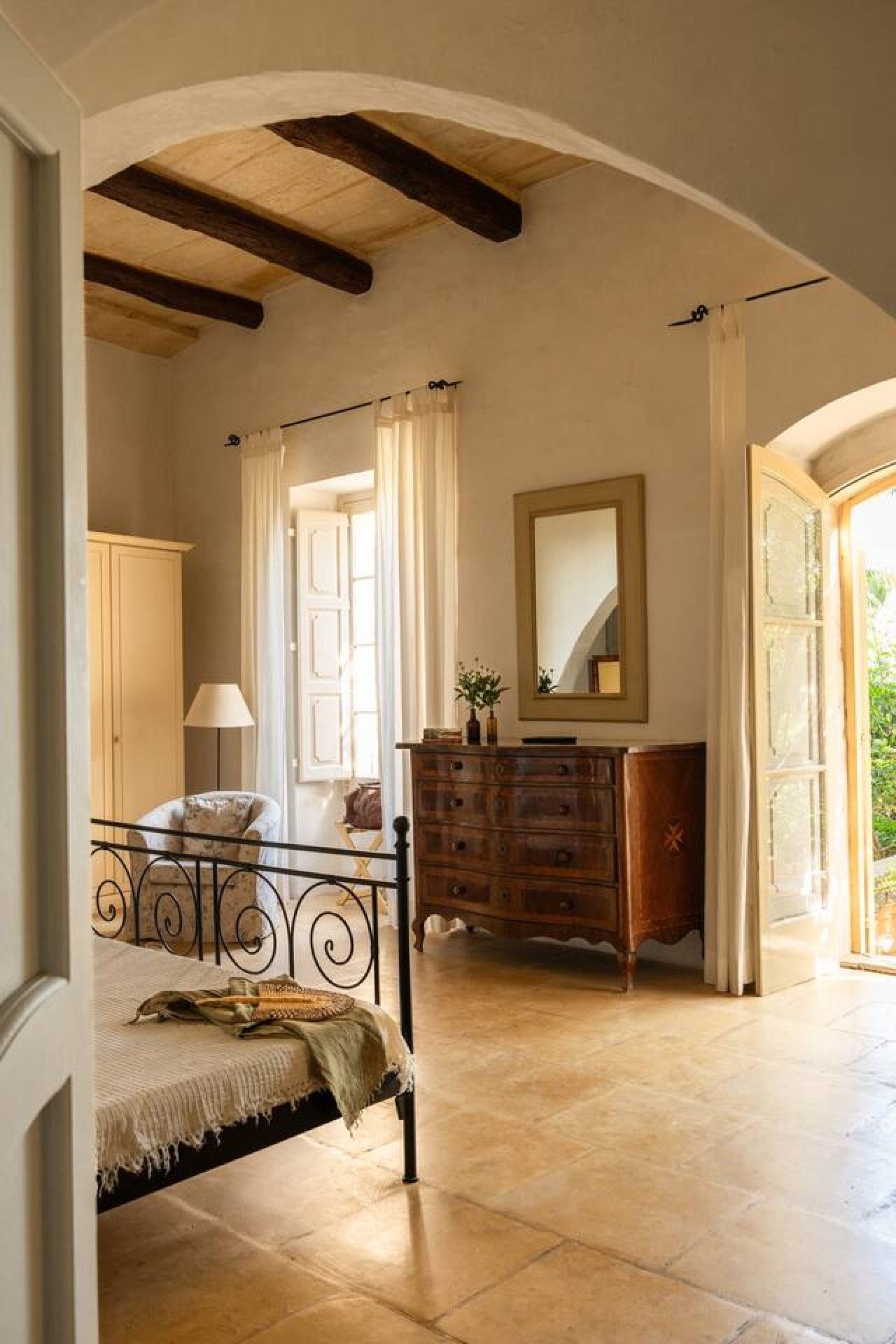 Une chambre à coucher avec un lit, une commode et un miroir. Il y a une fenêtre avec des rideaux et une vue sur la verdure à l’extérieur.