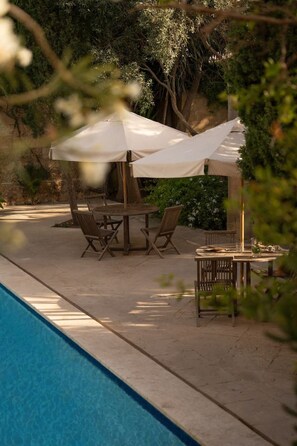 A poolside area with a table and chairs under a canopy, surrounded by greenery.