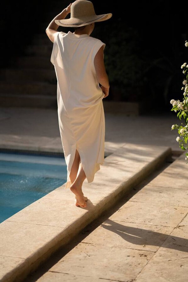 Une personne se promène au bord d’une piscine, vêtue d’une robe blanche et d’un chapeau, près de la piscine et de quelques plantes.