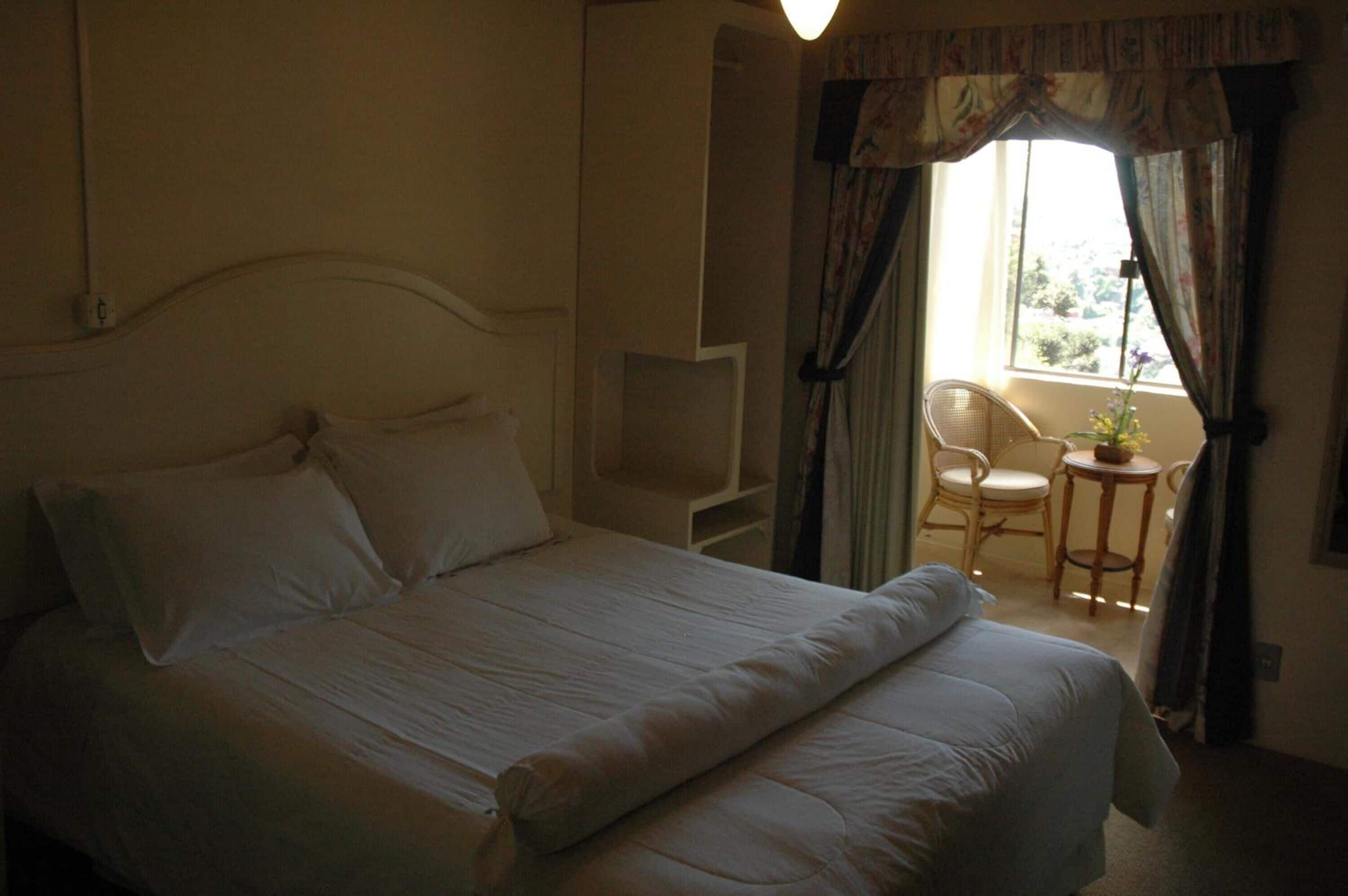 A neatly made bed with white linens in a room with a window, a chair, and a small table.