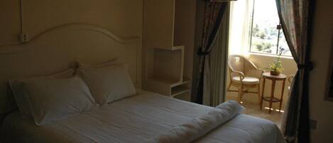 A neatly made bed with white linens in a room with a window, a chair, and a small table.