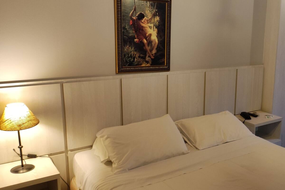 Una habitación de hotel con una cama, una mesita de noche con una lámpara, un cuadro en la pared, una silla y un armario.