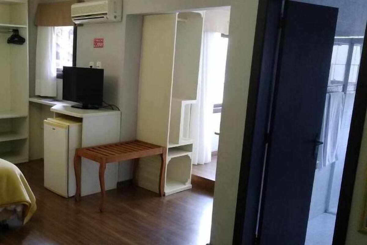 Habitación de hotel compacta con cama, escritorio, televisor y un pequeño taburete de madera.