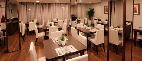 A hotel dining area with multiple tables and chairs, a reception desk, and a large mirror.