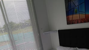 A neatly made bed with white linens and a folded towel, a window with blinds, and a wall-mounted shelf. - Apartamento Turístico Familiar (Montenegro)