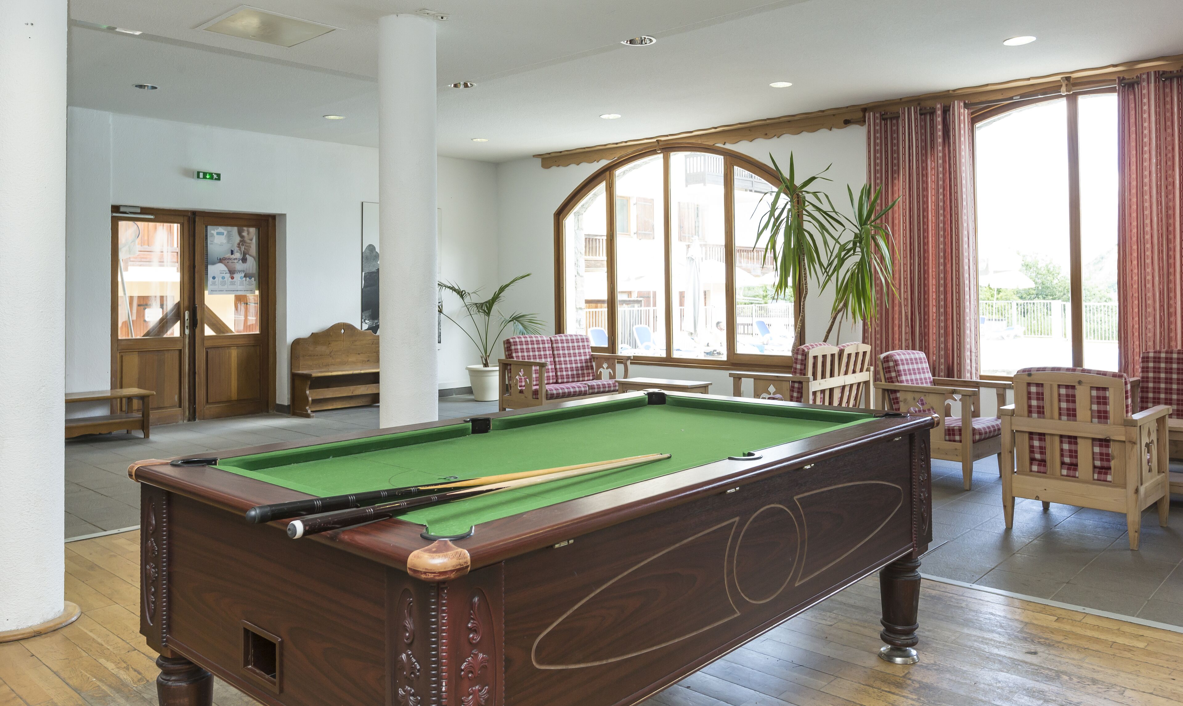 A pool table in a room with a large window, a bench, and a potted plant.