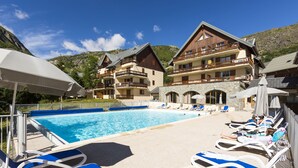 Un área junto a la piscina con sillones y sombrillas, piscina y un edificio con balcones.