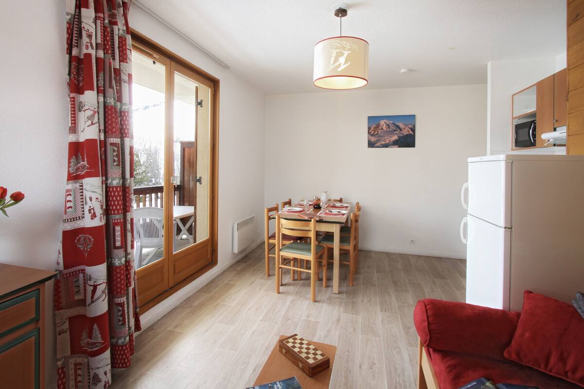 A room with a dining table, chairs, a red sofa, a wooden cabinet, a white refrigerator, and a window with a curtain.