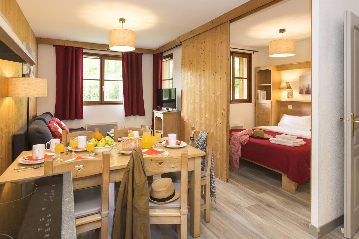 Une pièce chaleureuse avec une table en bois mise pour le petit-déjeuner, un lit recouvert d’une couverture rouge et un téléviseur posé sur un meuble en bois.