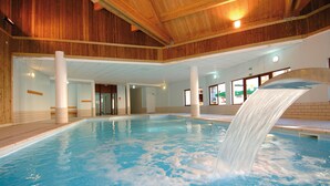 Piscina interna com cachoeira, teto de madeira e paredes revestidas de azulejo.