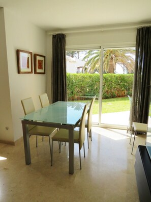 Zona pranzo con tavolo e sedie in vetro, una poltrona, televisione e vista su un giardino con palme.