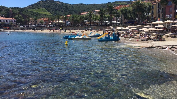 Una playa con agua cristalina, barcos y una colina con edificios al fondo.