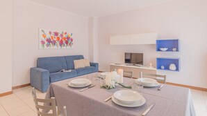 A dining area with a table set for four, a blue sofa, a TV, and decorative shelves. - Alexis House Varenna (Varenna)
