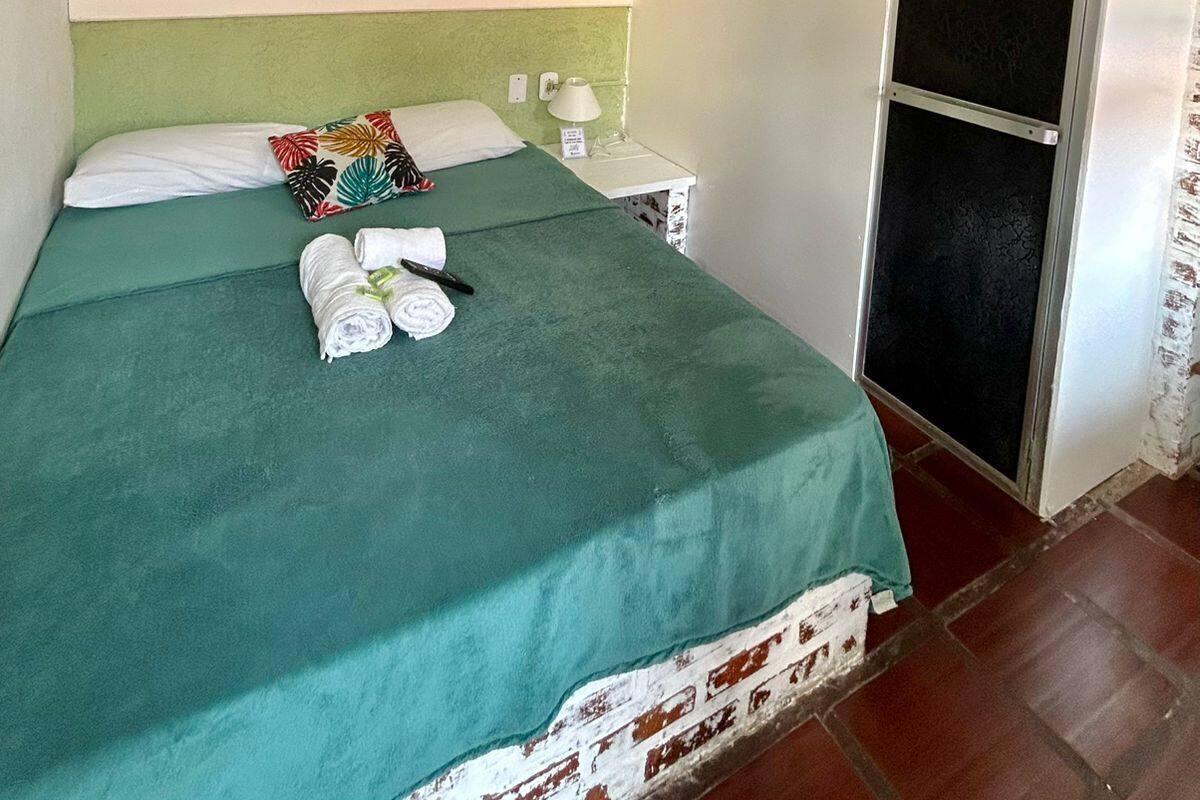 Une chambre avec un lit simple, une couvre-lit verte, des oreillers blancs, un ventilateur de plafond, une petite table de chevet avec une lampe et une fenêtre munie d’un moustiquaire métallique.