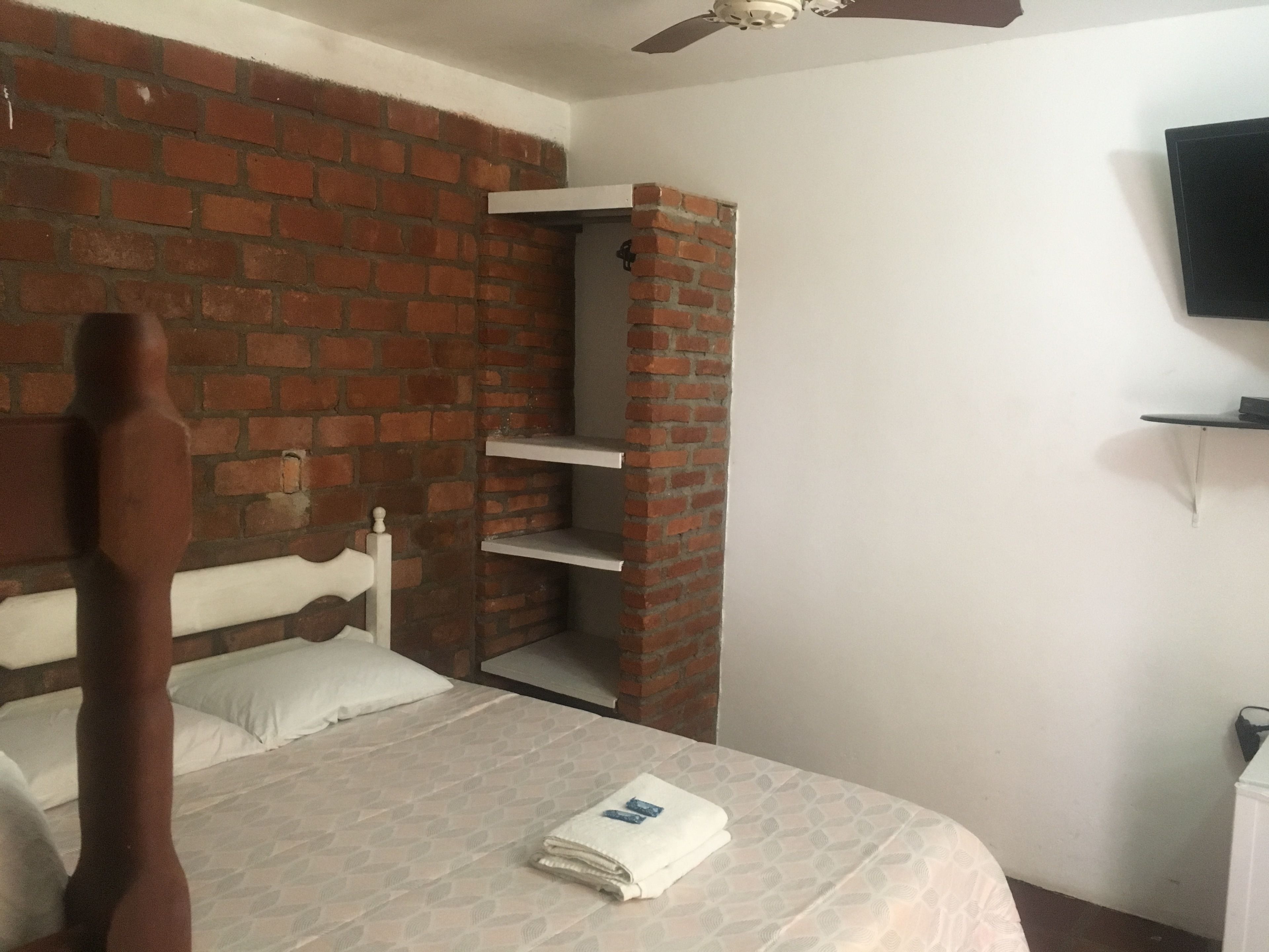 A bedroom with a brick wall, a wooden bed frame, a white headboard, a flat-screen TV mounted on the wall, and a small shelf unit.