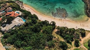 An aerial view of a coastal area with a sandy beach, clear turquoise waters, and a small island with boats. - Pousada Aldeia Buzios (Búzios)