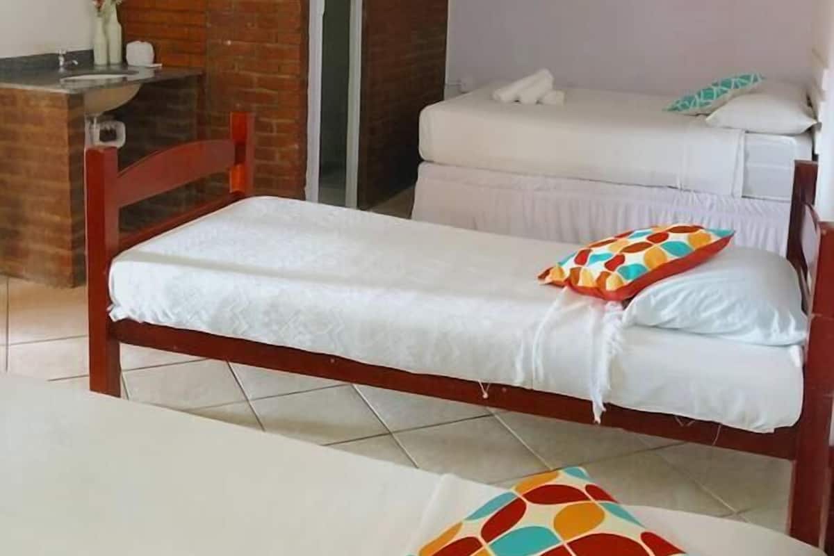 Une petite chambre avec un lit simple, un cadre de lit en bois, un matelas blanc et un oreiller coloré. On y trouve un petit bureau avec un miroir et un vase de fleurs. La chambre a un mur en briques et un ventilateur de plafond.