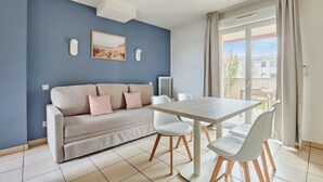 A modern living room with a sofa, dining table, and chairs. There is a painting on the wall and a window with curtains. - Résidence Odalys Le Mas des Flamants (Aigues-Mortes)
