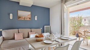 A modern living room with a sofa, a dining table with breakfast items, and a large window with a view of the outdoors. - Résidence Odalys Le Mas des Flamants (Aigues-Mortes)