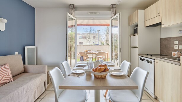 A modern kitchen with white cabinets, a dining table set for four, and a view of the outdoors. - Résidence Odalys Le Mas des Flamants (Aigues-Mortes)
