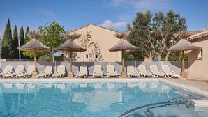 A swimming pool with lounge chairs and thatched umbrellas, surrounded by trees and a building. - Résidence Odalys Le Mas des Flamants (Aigues-Mortes)