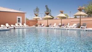 Una piscina con sillas de descanso y sombrillas, un edificio de tejado a la vista y un cielo azul despejado.