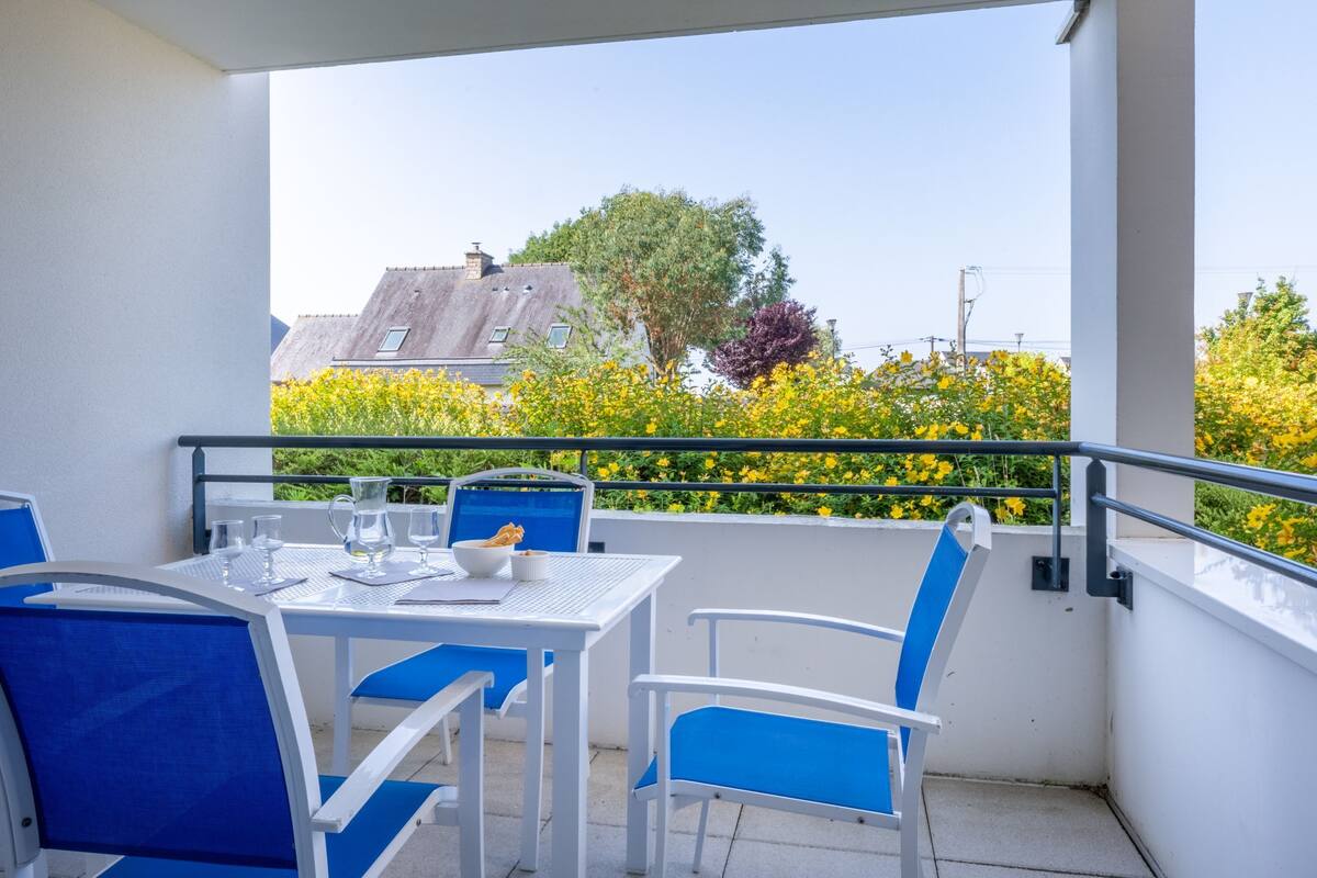 Een balkon met een tafel en stoelen, een hekwerk en uitzicht op gebouwen en bomen.