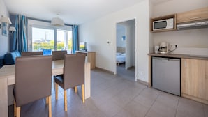 A modern kitchen with a microwave, coffee maker, and dishwasher. There is a dining area with a table and chairs, and a view of the outdoors through large windows. - Résidence Odalys Les Iles du Morbihan (Baden)